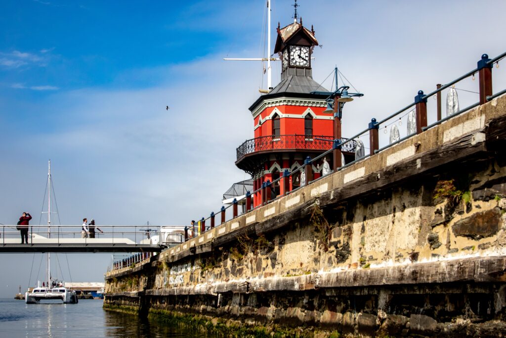 Roter Uhrturm an der V&A Waterfront in Kapstadt mit Hafenpromenade, Segelbooten und blauem Himmel