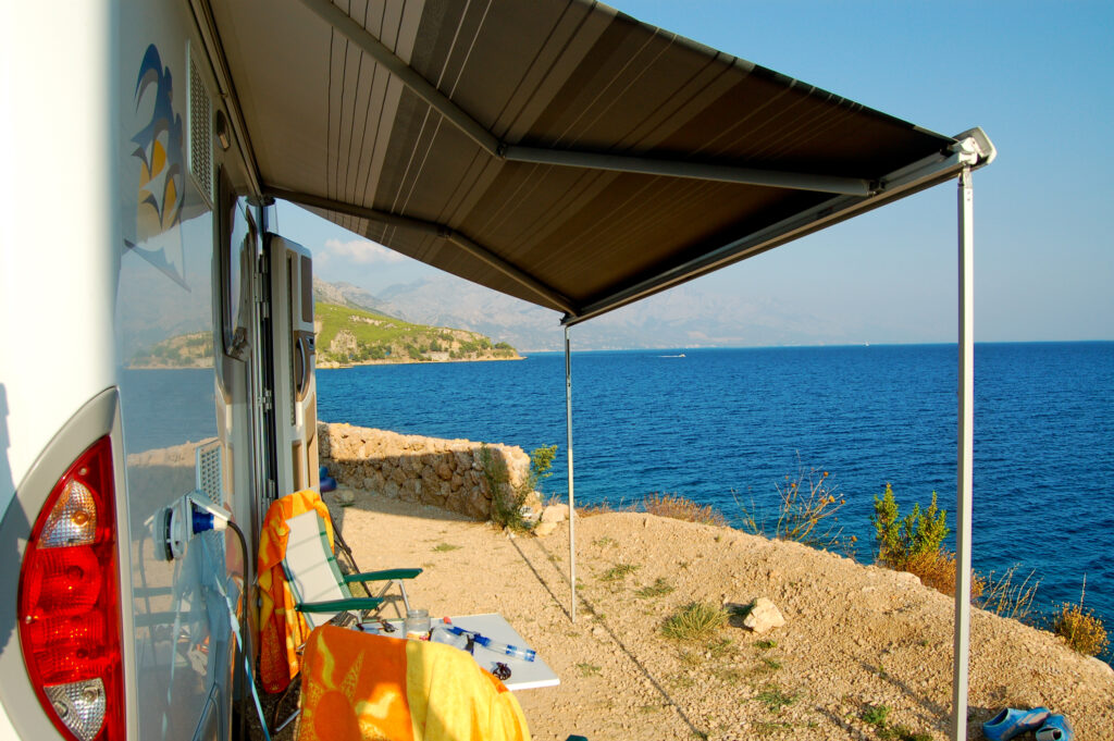 Wohnmobil mit ausgefahrener Markise und Campingstuhl direkt an der Küste mit Blick auf das Meer und die Berge